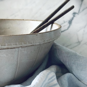 Puede incluir: Un cuenco de cerámica blanquecina con superficie texturizada y borde decorativo. Dos palillos de madera oscura descansan dentro del cuenco. El cuenco está sobre un paño de lino a rayas azules y blancas, sobre un fondo de mármol.