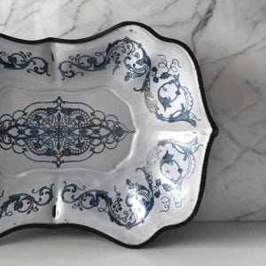 Puede incluir: Bandeja de esmalte blanco con borde negro y diseños florales azules ornamentados. La bandeja tiene un borde festoneado y un patrón decorativo central. Una pieza de servicio de estilo vintage.