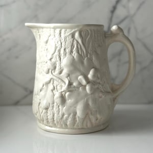 May include: A cream-colored ceramic pitcher featuring a raised, textured relief pattern of oak leaves and acorns across the exterior surface. The vessel has a curved handle on the right side and a wide pouring spout at the top rim.