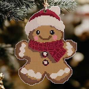 Peut inclure: Une décoration de Noël en forme d'homme en pain d'épices avec un chapeau rouge et blanc et une écharpe rouge. L'homme en pain d'épices sourit et a du glaçage blanc sur ses bras et ses jambes.