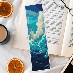 Op de afbeelding: Een boekenlegger met een gepixeld oceaanontwerp in blauw- en wittinten ligt op een open boek. De scène bevat gedroogde sinaasappelschijfjes, een zwarte mok en een bril, wat een gezellige leessfeer creëert.