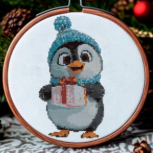 Op de afbeelding: Kruissteekborduurwerk van een pinguïn met een blauwe muts en sjaal, die een cadeau vasthoudt. De pinguïn zit in een houten borduurring, met kerstversieringen op de achtergrond.
