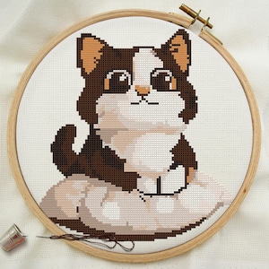 Op de afbeelding: Een kruissteekborduursel van een bruine, witte en oranje kat met grote ogen die op een wit kussen zit. Het borduursel is ingelijst in een houten ring.