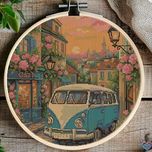 Könnte beinhalten: Ein blauer und weißer Vintage-Campingbus, der auf einer Kopfsteinpflasterstraße in einer europäischen Stadt geparkt ist. Die Straße ist gesäumt von Gebäuden und Blumen. Der Himmel ist ein leuchtend orangefarbener und rosafarbener Sonnenuntergang.