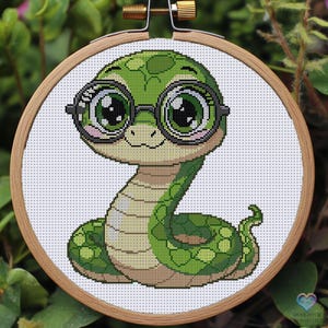 Könnte beinhalten: Kreuzstich-Stickerei einer Cartoon-Schlange mit Brille. Die Schlange ist grün mit dunkelgrünen Flecken und einem hellbraunen Bauch. Die Stickerei befindet sich in einem Holzreifen.