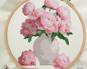 May include: A cross-stitch embroidery of a bouquet of pink peonies in a white vase. The peonies are arranged in a cluster with green leaves and stems. The vase is a simple, round shape with a white background.