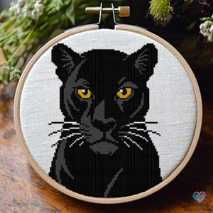 Op de afbeelding: Een zwarte panter met gele ogen, die rechtstreeks naar de kijker kijkt. De panter is geborduurd op een witte stof, die gespannen is over een houten borduurring.
