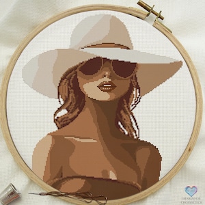 Pode incluir: Um padrão de ponto cruz de uma mulher usando um grande chapéu de sol branco e óculos de sol. A mulher tem cabelo castanho comprido e está usando um top marrom.