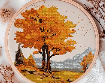 May include: A cross-stitch embroidery of a fall landscape with a large orange and yellow tree in the center. The tree is surrounded by smaller trees and bushes, and the background is a light blue sky with white clouds.