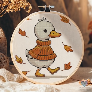 May include: A cross-stitch embroidery hoop showing a duck in an orange sweater, walking amongst falling autumn leaves. The duck is white with an orange beak and feet. The text "DESIGNFOR CROSSSTITCH" is visible in the lower right corner.