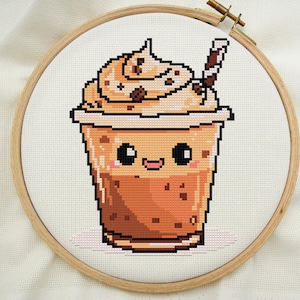 Peut inclure: Un motif de point de croix représentant un café glacé mignon avec une paille et de la crème fouettée. Le café a un visage souriant avec de grands yeux.