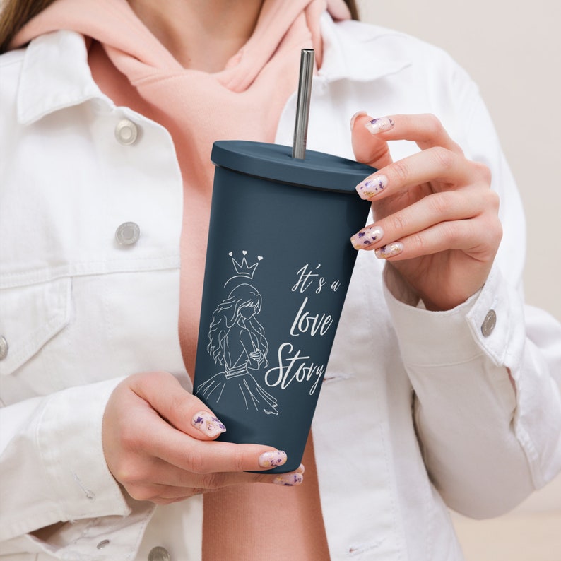 May include: A navy blue tumbler with a silver straw. The tumbler features a white illustration of a woman wearing a crown, with the text "It's a love Story". The tumbler is held by a person wearing a white jacket.