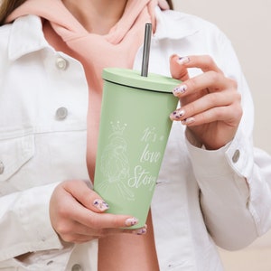May include: A light green tumbler with a silver straw and a black lid. The tumbler features a white illustration of a princess and the text "It's a love Story". The tumbler is held by a person wearing a white jacket.