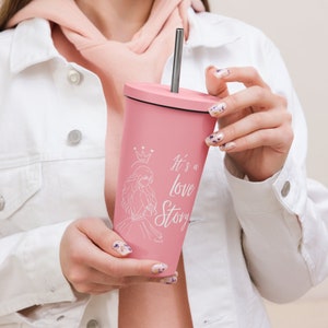 May include: A pink tumbler with a black lid and a silver straw. The tumbler features a white illustration of a princess and the text "It's a love Story". The tumbler is held by a person wearing a white jacket.