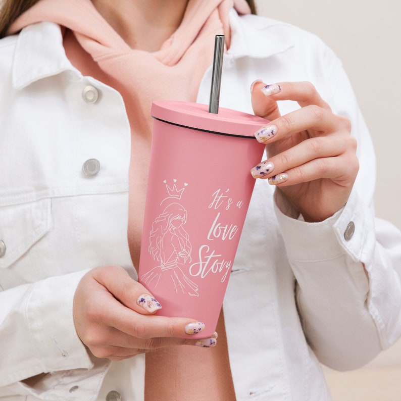 May include: A pink tumbler with a black lid and a silver straw. The tumbler features a white illustration of a princess and the text "It's a love Story". The tumbler is held by a person wearing a white jacket.