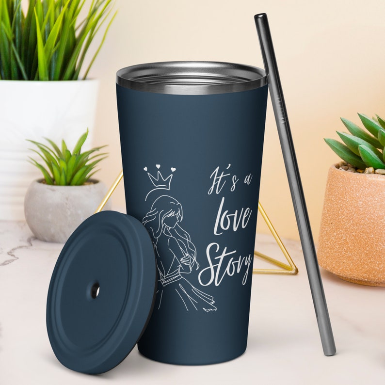 May include: A navy blue travel tumbler with a silver rim, lid, and straw. The tumbler features a white illustration of a woman with a crown and the text "It's a love Story". The lid is off to the side.