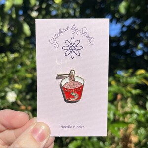 May include: A pink card with the text "Stitched by Sophie" and a flower design. A red and white enamel pin in the shape of a ramen bowl is attached to the card. The pin has the word "NOODLES" on it. The card also says "Needle Minder".
