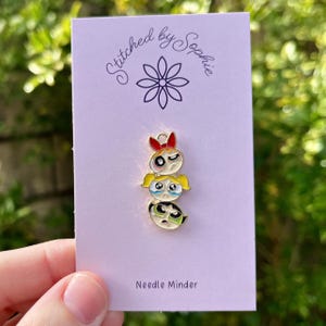 Peut inclure: Un aimant à aiguille doré représentant trois personnages de dessins animés empilés avec des détails rouges, jaunes et verts. L'aimant à aiguille est sur une carte violet clair avec le texte "Stitched by Sophie" et "Needle Minder".