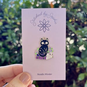 May include: Black cat needle minder with gold accents sitting on a stack of purple books with flowers. The needle minder is labelled "Stitched by Sophie" and "Needle Minder".