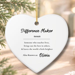 Könnte beinhalten: Ein weißes, herzförmiges Keramikornament mit einer goldenen Aufhängeschnur. Der Text lautet: Difference Maker, noun, Someone who touches lives, brings out the best in others, & leaves the world a little brighter, Also Known as: Olivia.