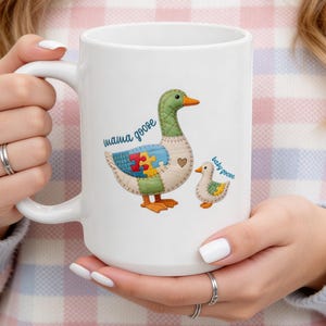 Op de afbeelding: Witte keramische mok met een illustratie van een moedergans en een babygans. De mama-gans heeft een patchwork-ontwerp met een puzzelstukje en de baby-gans is kleiner. Tekst op de mok luidt "mama goose" en "baby goose".