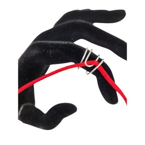 May include: A silver ring with three loops, designed to hold a cord or string. The ring is shown on a black velvet hand model with a red cord threaded through the loops.