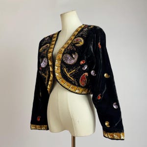 Peut inclure: Boléro en velours noir avec garniture de sequins dorés et broderies ornées. La veste présente des motifs cachemire et floraux colorés, avec une coupe courte et des manches longues. Une pièce vintage.