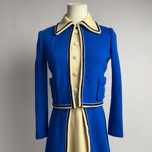 Könnte beinhalten: Ein Vintage-Rockanzug in Blau mit cremefarbenem Oberteil und Rock. Die Jacke hat einen cremefarbenen Kragen mit schwarzem Besatz, passend zu den Taschen der Jacke und den Kanten des Rocks. Der Anzug hat cremefarbene Knöpfe und wird auf einer Schaufenquin präsentiert.