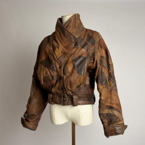 Op de afbeelding: Bruine leren jas met een patchwork ontwerp. De jas heeft een hoge kraag, een knoopsluiting en een riem in de taille. De jas wordt op een mannequin gepresenteerd.