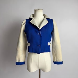 Könnte beinhalten: Eine kurze Jacke mit blauem Vorderteil und cremefarbenen Ärmeln. Sie hat einen weißen Kragen, blaue Knöpfe und kleine silberfarbene Schnallen. Die Ärmel haben blaue Manschetten. Die Jacke wird auf einer Schaufensterpuppe präsentiert.