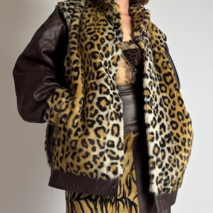 Pode incluir: Um casaco com painel frontal de pele sintética com estampado de leopardo e mangas de couro castanho escuro. A peça tem gola alta, fecho frontal com botões e bolsos laterais. Usado sobre um top de leopardo e uma saia castanha com riscas de tigre.