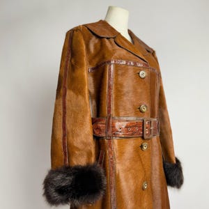 Pode incluir: Um casaco castanho vintage com gola e punhos de pele. O casaco apresenta um cinto com fivela e costuras decorativas. O casaco está abotoado e tem um design clássico e intemporal.