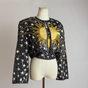 Puede incluir: Chaqueta bolero negra con cuentas con un diseño de sol y estrellas. La chaqueta presenta un gran diseño de sol en oro y lentejuelas iridiscentes en la parte delantera. Las mangas y el cuerpo están cubiertos de lentejuelas blancas en forma de estrella.
