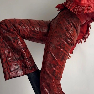 Op de afbeelding: Rode broek met slangenpatroon en een bijpassende rode leren jas met franjes. De broek heeft een uitlopende zoom en wordt gecombineerd met zwarte laarzen. Een opvallende mode-ensemble.