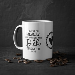 Könnte beinhalten: Weiße Keramiktasse mit einem schwarzen Design, auf dem "Mögest du immer das machen, was Dich glücklich macht" mit einem Herzen und einem Kranz drumherum steht.