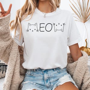 Könnte beinhalten: Weißes T-Shirt mit einer schwarzen Grafik von zwei Comic-Katzen und dem Wort "MEOW" dazwischen.