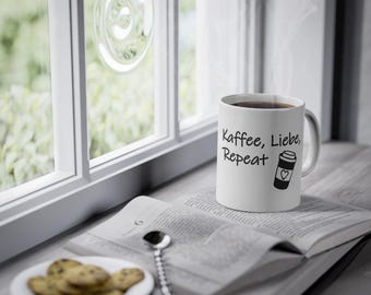 May include: A white ceramic mug featuring the black printed text Kaffee, Liebe, Repeat and a graphic of a coffee cup with a heart icon. The mug is filled with a dark liquid and sits next to an open book and a small plate of chocolate chip cookies.