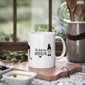 Könnte beinhalten: Weiße Keramiktasse mit einer schwarzen Grafik eines Gnom und dem deutschen Text "Du bist mir Wicht(el)ig" und einem Herz. Die Tasse steht auf einem Holzschneidebrett, mit Oliven und einem Fenster im Hintergrund.