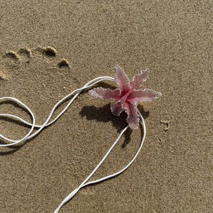 May include: A pink beaded flower with a white string lies on a sandy beach. The flower has six petals and a delicate, textured appearance. The white string is looped and extends across the sand.