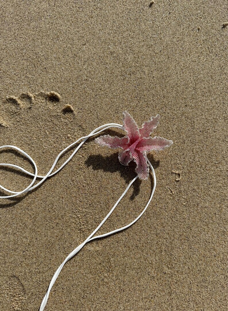 May include: A pink beaded flower with a white string lies on a sandy beach. The flower has six petals and a delicate, textured appearance. The white string is looped and extends across the sand.