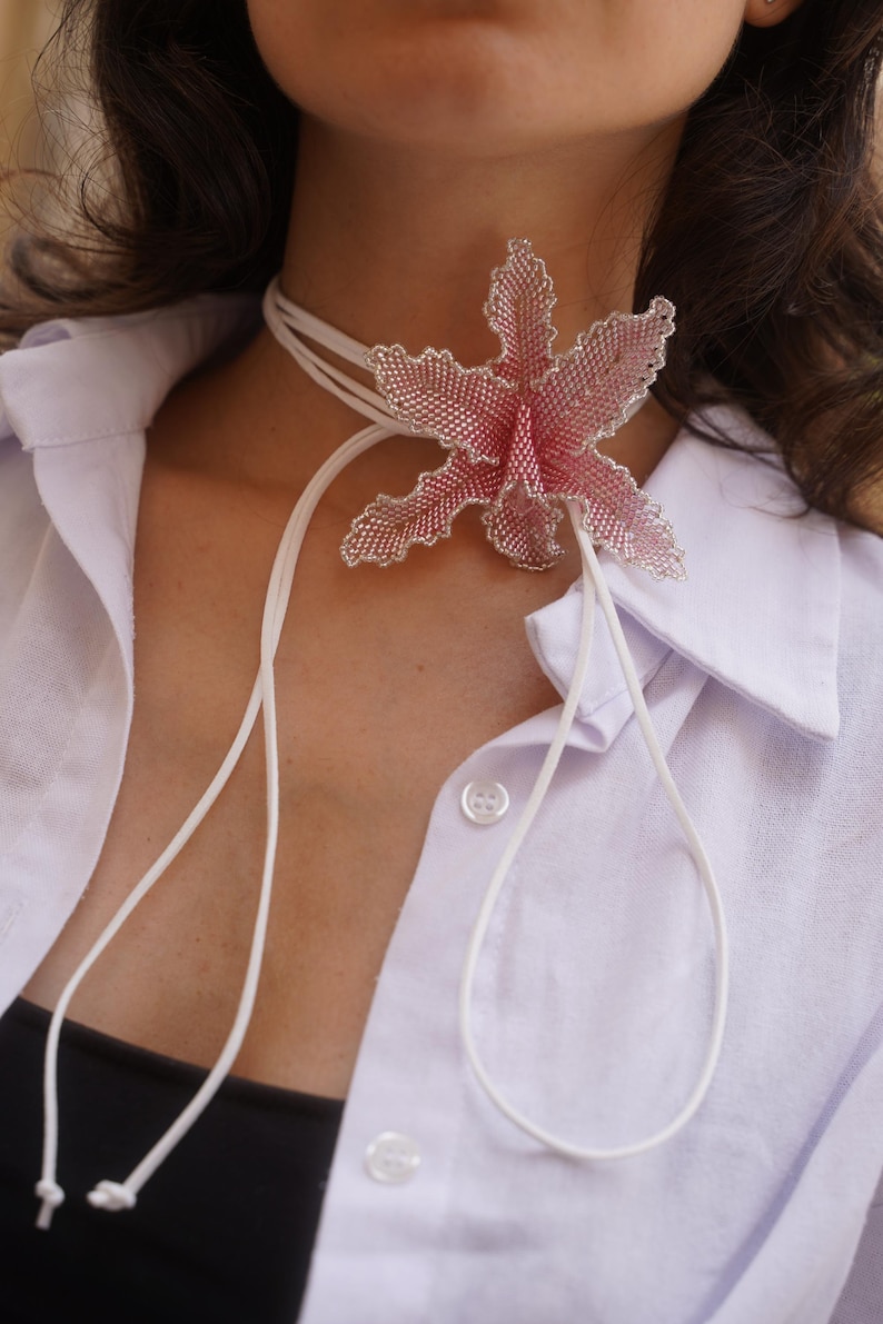 May include: A beaded flower choker necklace with a pink and silver design. The flower is the focal point, attached to a white cord that ties in the back. The necklace is worn over a white button-down shirt.