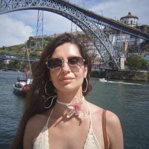 May include: A woman wearing sunglasses, a white crochet halter top, and a floral choker stands in front of a large metal bridge over a river. The background includes buildings and boats under a blue sky.