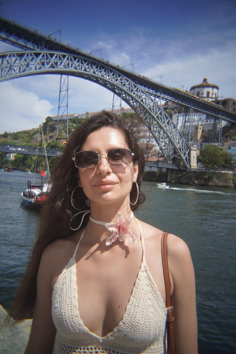 May include: A woman wearing sunglasses, a white crochet halter top, and a floral choker stands in front of a large metal bridge over a river. The background includes buildings and boats under a blue sky.
