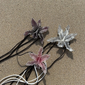 May include: Three beaded flower pendants in shades of pink, gray, and purple, each with a black or white cord. The flowers are detailed with small beads, and the cords are lying on a sandy surface.