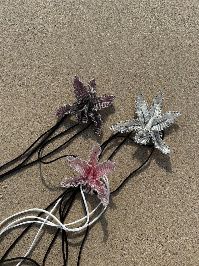 May include: Three beaded flower pendants in shades of pink, gray, and purple, each with a black or white cord. The flowers are detailed with small beads, and the cords are lying on a sandy surface.