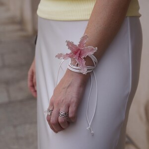 May include: A beaded flower bracelet in shades of pink and silver, wrapped around the wrist with white cords. The bracelet is the focal point, complemented by silver rings. The person is wearing a yellow top and white skirt.
