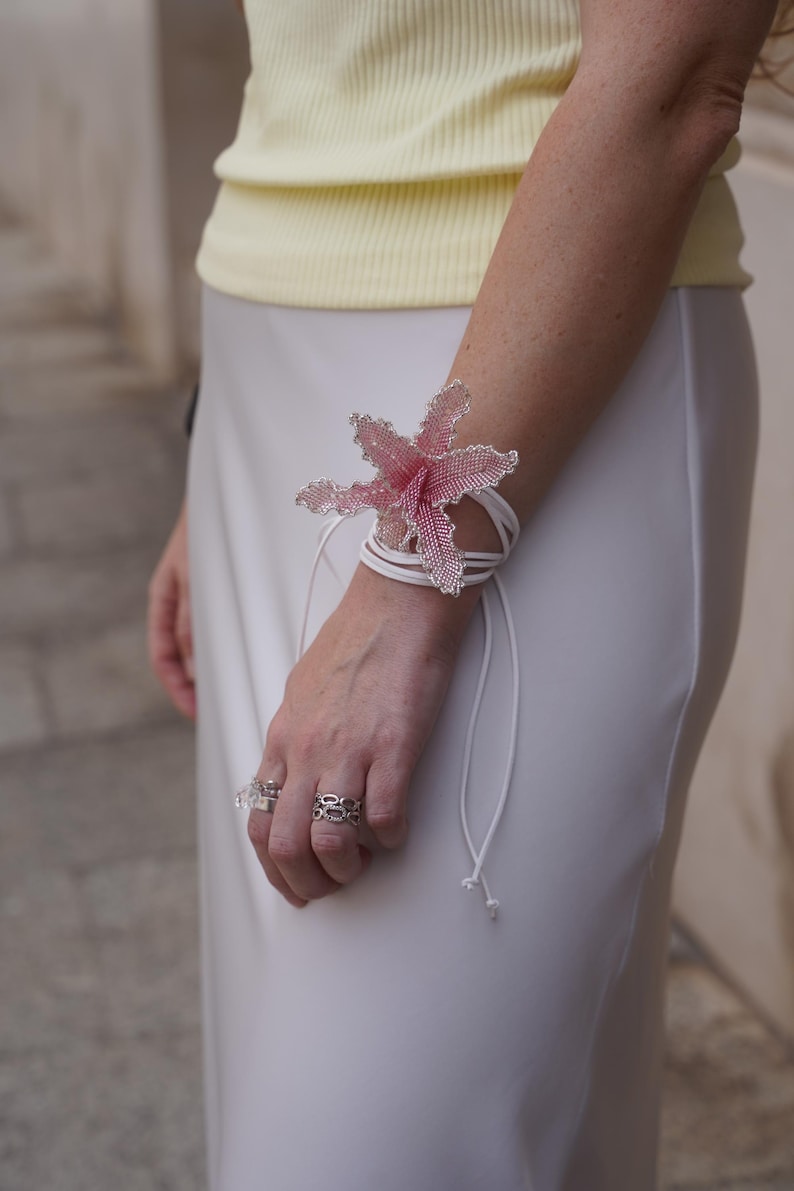 May include: A beaded flower bracelet in shades of pink and silver, wrapped around the wrist with white cords. The bracelet is the focal point, complemented by silver rings. The person is wearing a yellow top and white skirt.