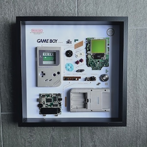 Könnte beinhalten: Gerahmtes Kunstwerk mit einer zerlegten Nintendo Game Boy von 1989. Die graue Konsole ist zerlegt, mit Komponenten wie Bildschirm, Knöpfen und Leiterplatten. Der Text "GAME BOY" und "Nintendo" sind sichtbar.