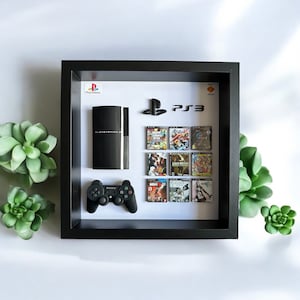 Op de afbeelding: Een zwarte, ingelijste schaduwdoos met een witte achtergrond. De schaduwdoos bevat een zwarte PlayStation 3 console, een zwarte controller en negen videospelcassettes. De videospelcassettes zijn gerangschikt in drie rijen van drie. De tekst "PlayStation" en "PS3" is zichtbaar in de afbeelding.