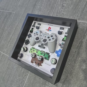 Puede incluir: Una exhibición enmarcada que presenta un mando de PlayStation One desarmado. El mando gris está centrado, rodeado de componentes etiquetados y el logotipo de Sony. El marco gris oscuro contrasta con el fondo de baldosas grises.